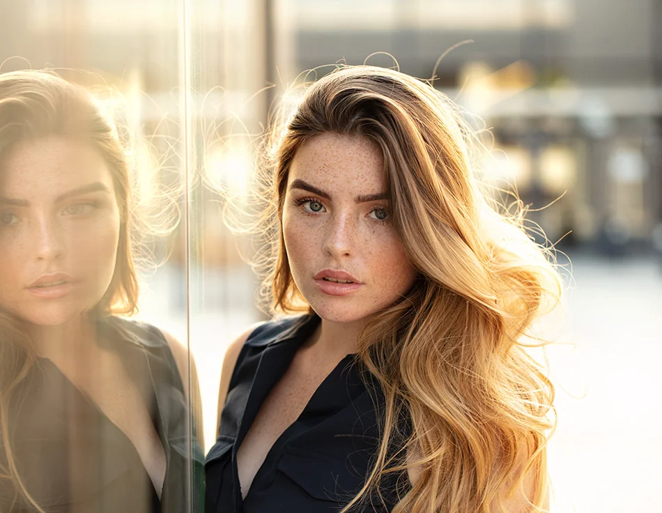 A woman with long, flowing hair and natural freckles gazes directly into the camera with a strong, confident expression, her reflection visible in a glass surface beside her - Lip Fillers in Houston, TX