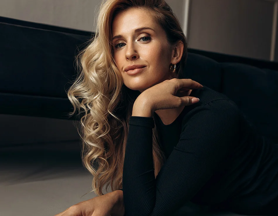 A woman with blonde, wavy hair, wearing a black top, lying down in a relaxed pose, looking confidently at the camera - Reuivion Skin Tightening in Houston, TX